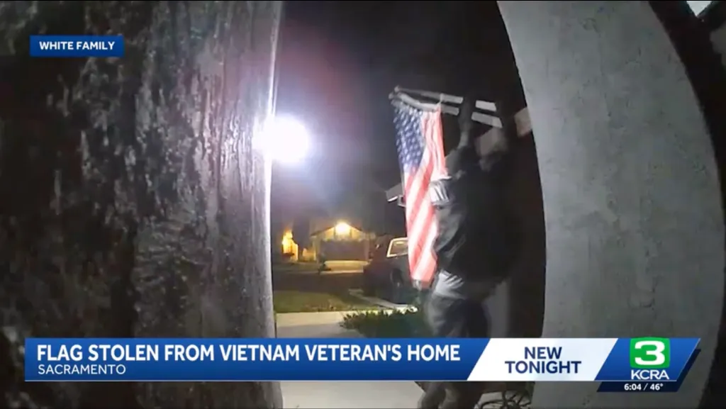 A person wearing a dark top and light pants taking an American flag from a pole next to a brightly lit porch at night.